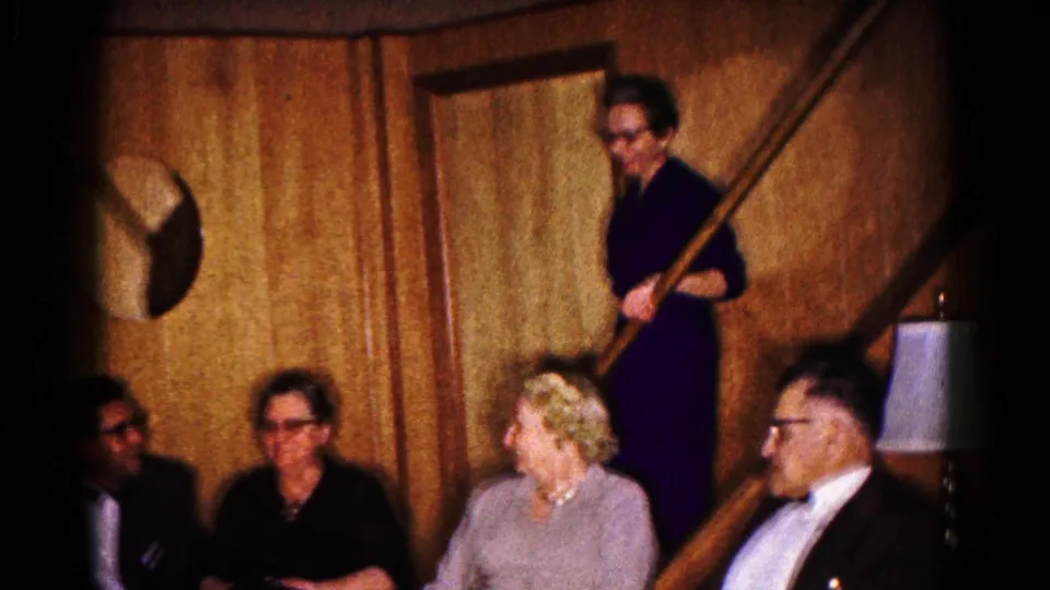 1961 Hagerstown home-movie frame showing adults in formal clothing seated below a staircase as a woman stands behind the banister.