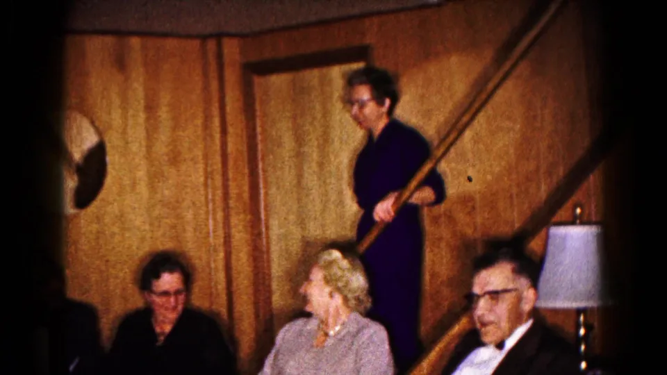1961 Hagerstown frame showing a woman holding the staircase banister above seated adults in a warm, wood-paneled private room.