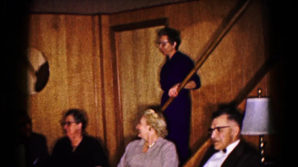 1961 Hagerstown home-movie frame showing older adults seated in a wood-paneled basement while a woman stands on the staircase.