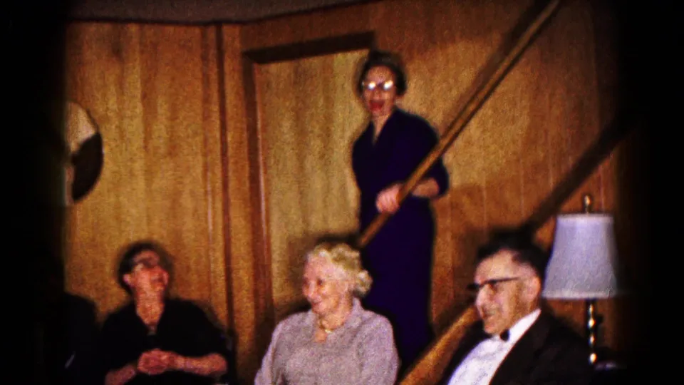 Older guests in formal attire sit near a staircase in a 1961 Hagerstown private interior, with one woman standing above them.