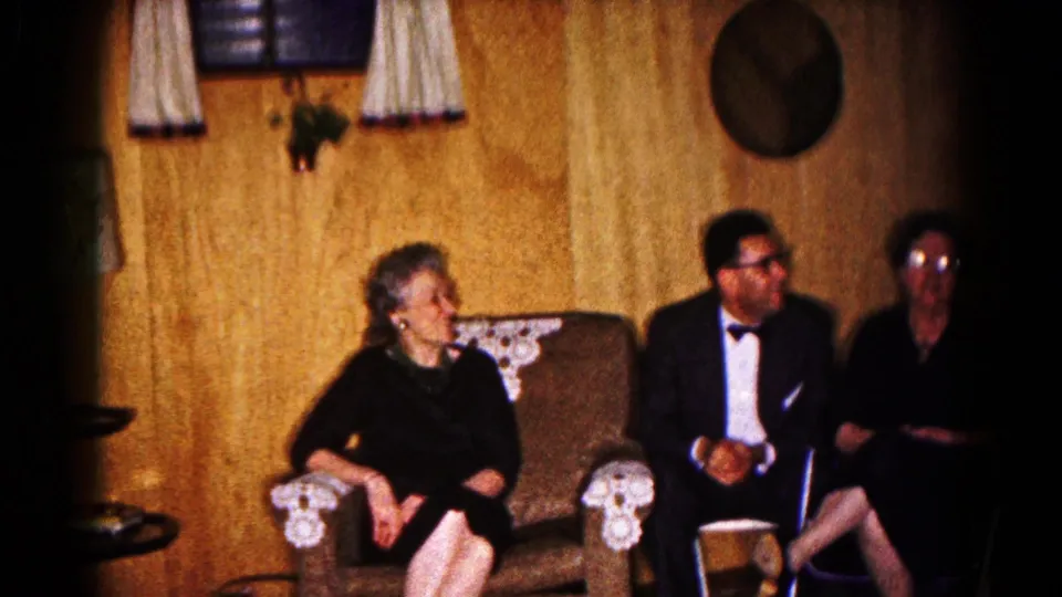Older guests in formal wear sit in a 1961 Hagerstown wood-paneled basement with curtains, wall decor, and upholstered chairs.