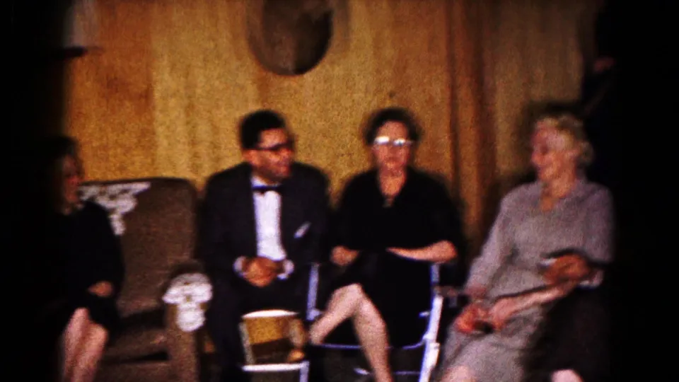Formally dressed adults sit and converse in a wood-paneled Hagerstown basement in 1961 home-movie footage.