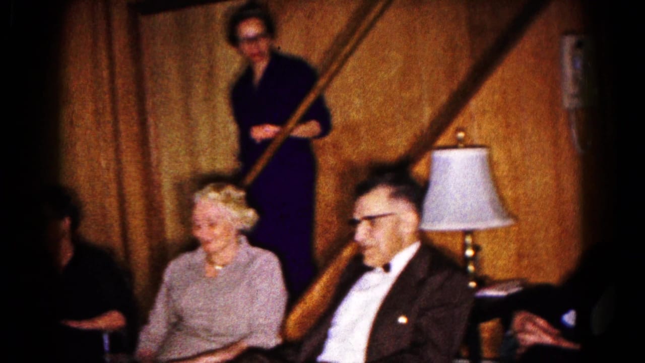 Adults sit near a staircase, with a woman standing in the background and a lamp nearby.