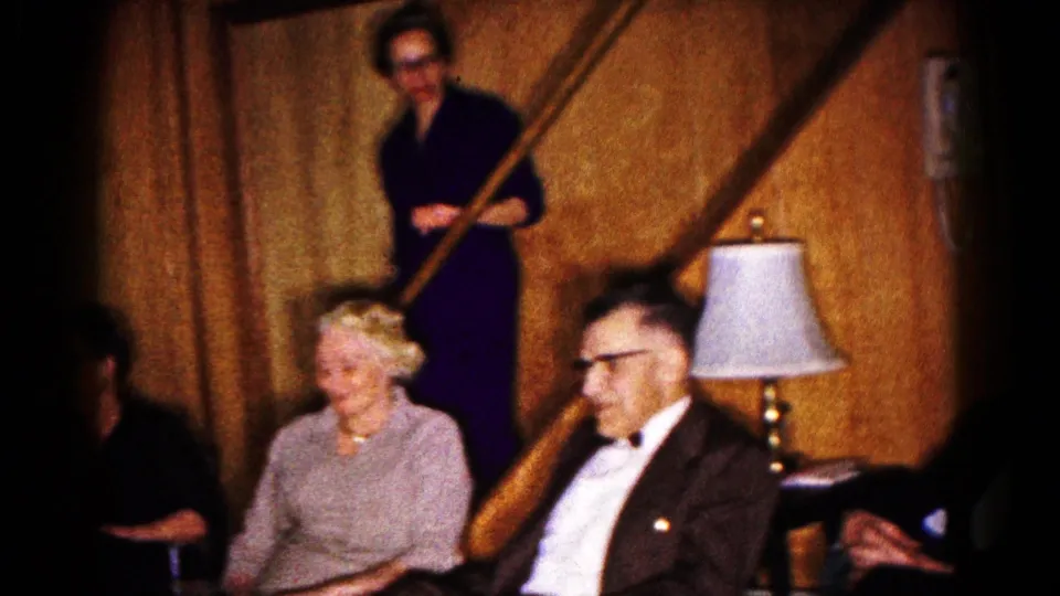 Adults in formal clothes sit near a basement staircase as a woman stands behind them in 1961 Hagerstown home-movie footage.