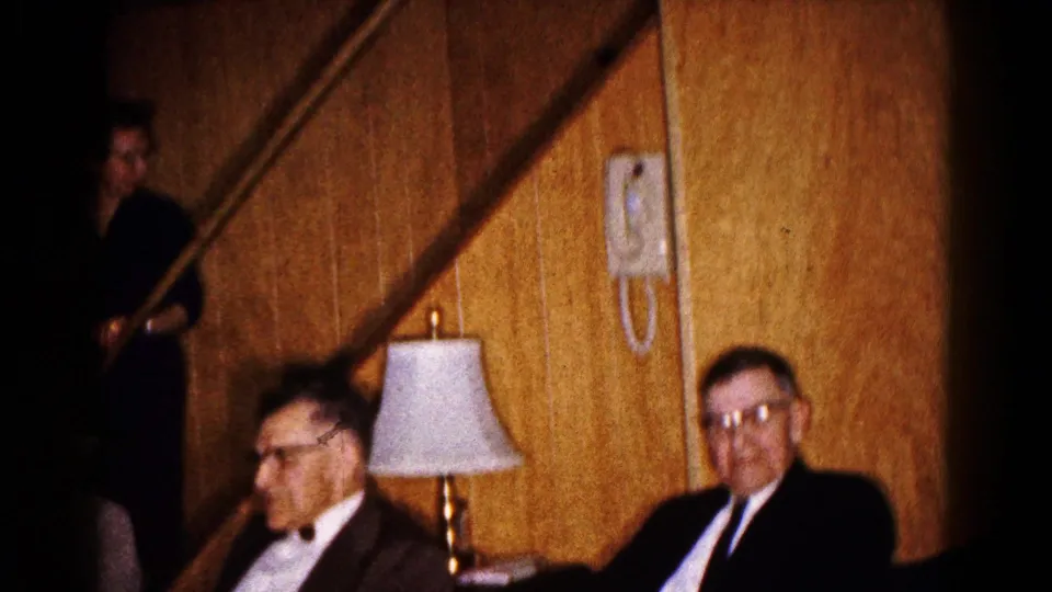 Formally dressed men sit in a paneled basement near a lamp and staircase during a 1961 Hagerstown home movie.