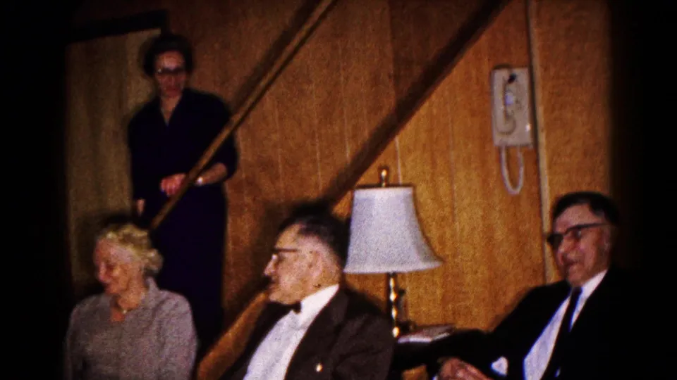 1961 Hagerstown home-movie frame showing older adults in formal clothing seated in a wood-paneled living room, with a woman by the stairs.