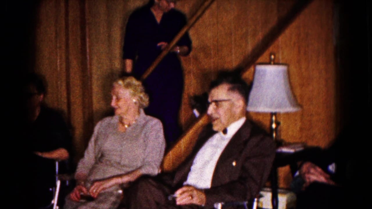 Seated older adults in formal clothing with a staircase and lamp behind them.