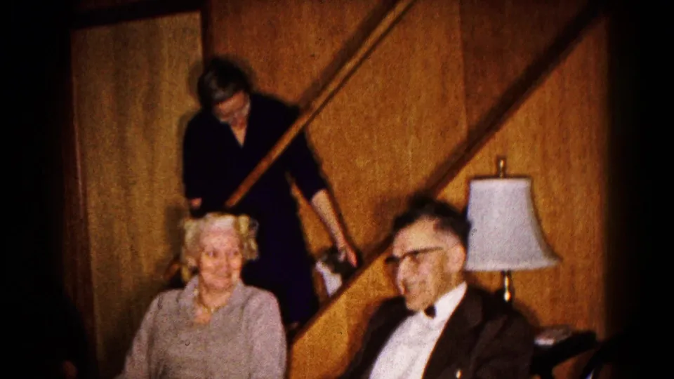 Home-movie frame of a woman seated near a man in a suit, with another woman standing by the staircase in a wood-paneled room.