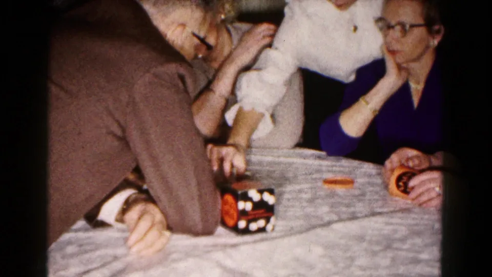 Close view of adults around a table during a dice game, with hands, dice, a dice cup, and seated figures visible in a private home.