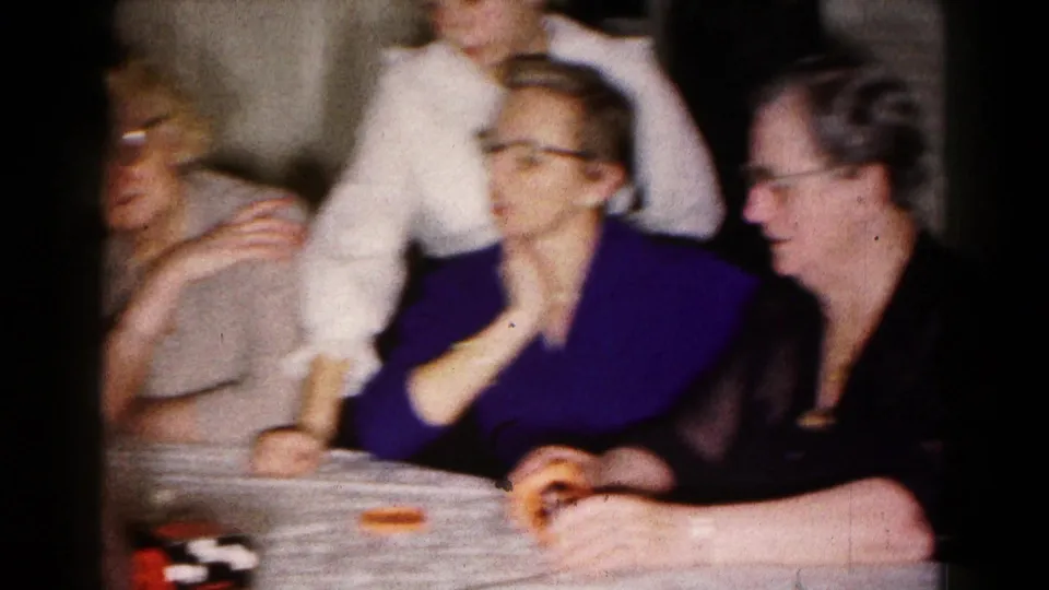 A home-movie frame shows adults seated at a Hagerstown kitchen table during a 1961 board or tabletop game.