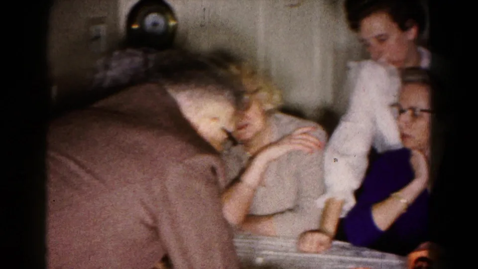 Adults cluster around a kitchen table in 1961 Hagerstown, framed softly on 8mm film during a tabletop game.