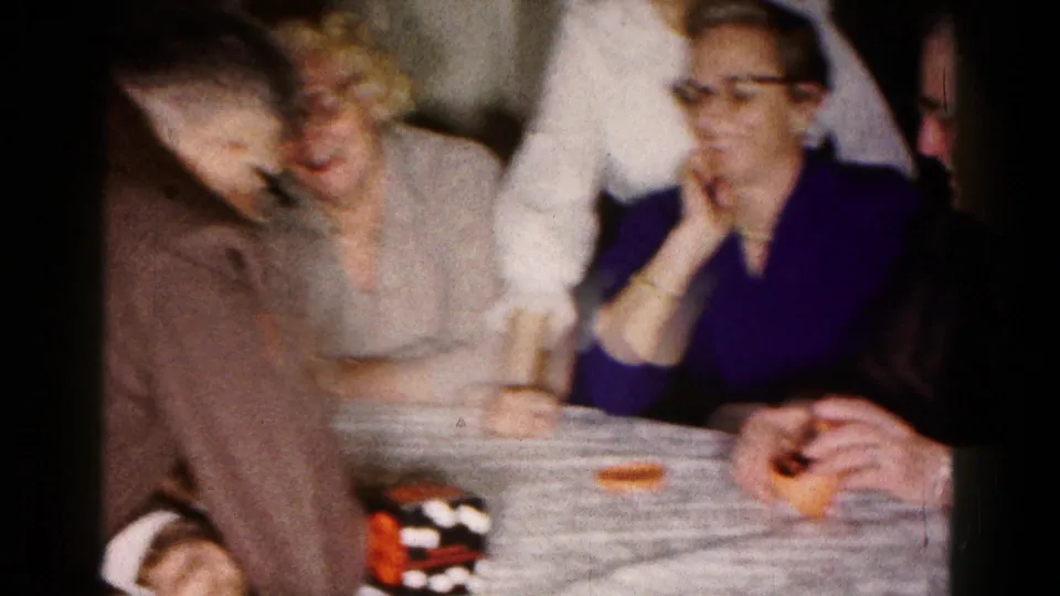 In the Hagerstown 1961 home movie, older women and a leaning man sit around the table during an indoor dice game.