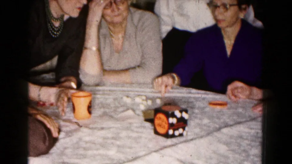 In a 1961 Hagerstown home-movie frame, adult women sit around a table covered with a light tablecloth while playing a dice game.