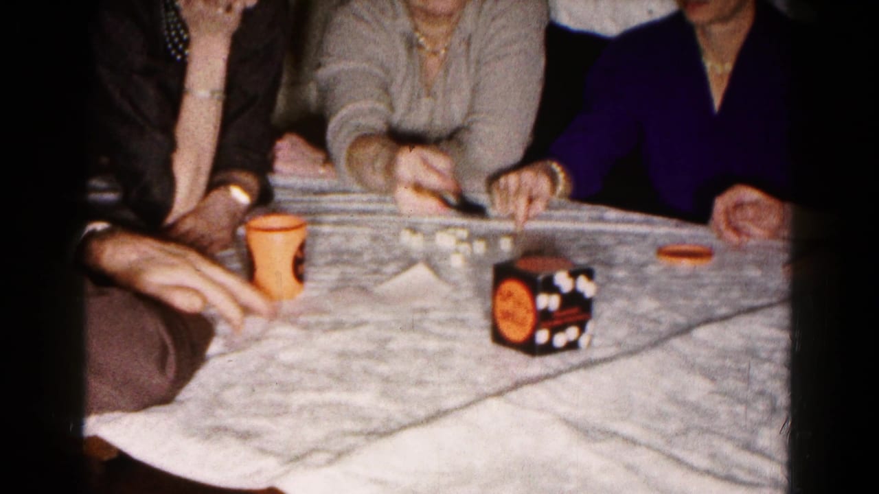 Hands and seated women around a table with dice, an orange cup, and a game cube.