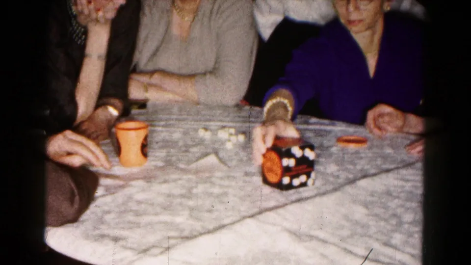 During the 1961 indoor game, a woman in a purple top reaches toward the game cube while others watch from around the table.