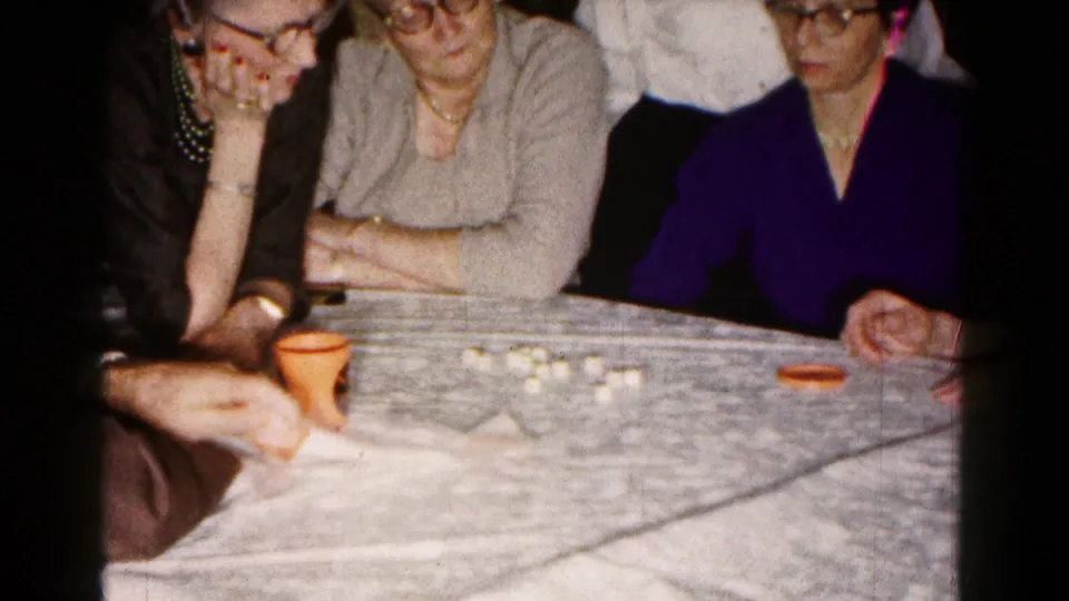 Three adult women, including one in a purple top and glasses, sit around a Hagerstown table with dice and an orange cup visible.