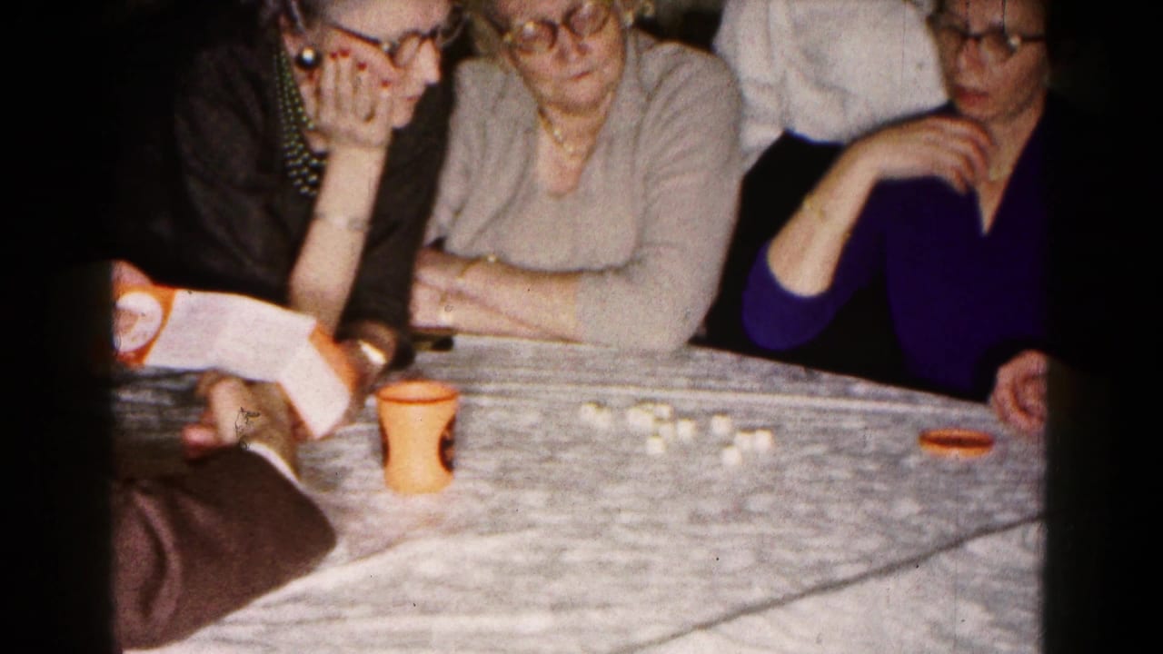 Three women sit at a table while a man holds a booklet near dice or game pieces.