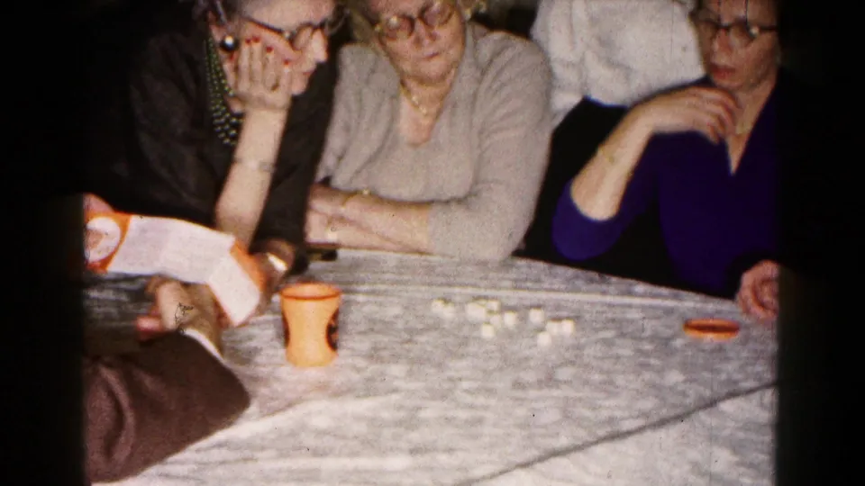 In a 1961 Hagerstown home-movie frame, three women watch as a man in a suit holds instructions beside small dice or game pieces on a table.