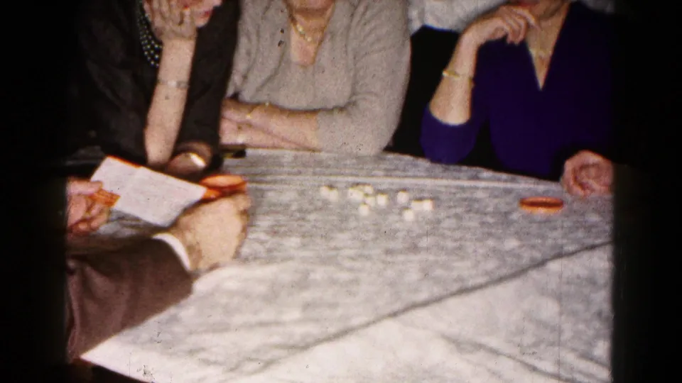 The home-movie frame shows women seated close together while a suited man handles instructions beside small white pieces and orange tableware.