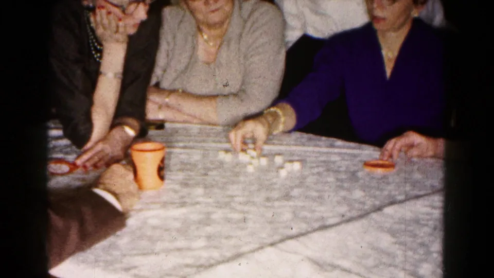 A woman in a purple top reaches toward the dice or pieces as the group continues the tabletop game in the 1961 Hagerstown home interior.