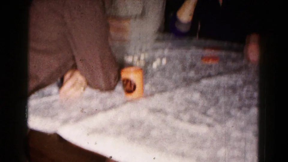 A soft 8mm close view shows a tablecloth, a seated adult's hand, and a cup during the indoor gathering.