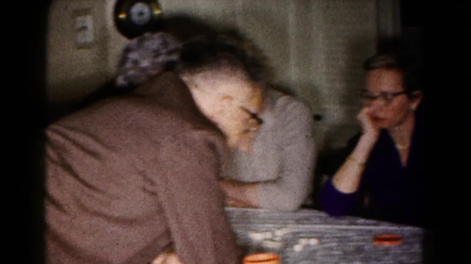 Adults sit around a dining table in the 1961 Hagerstown home-movie scene, with dishes and cups visible in soft 8mm focus.