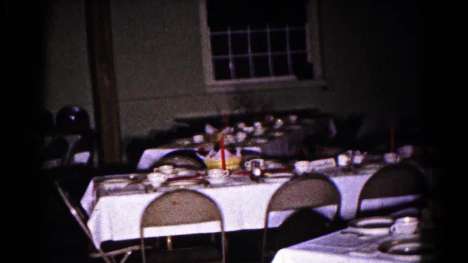 The room opens to a wider view of the table setting, folding chairs, dishes, and window in the 1961 Hagerstown indoor gathering.
