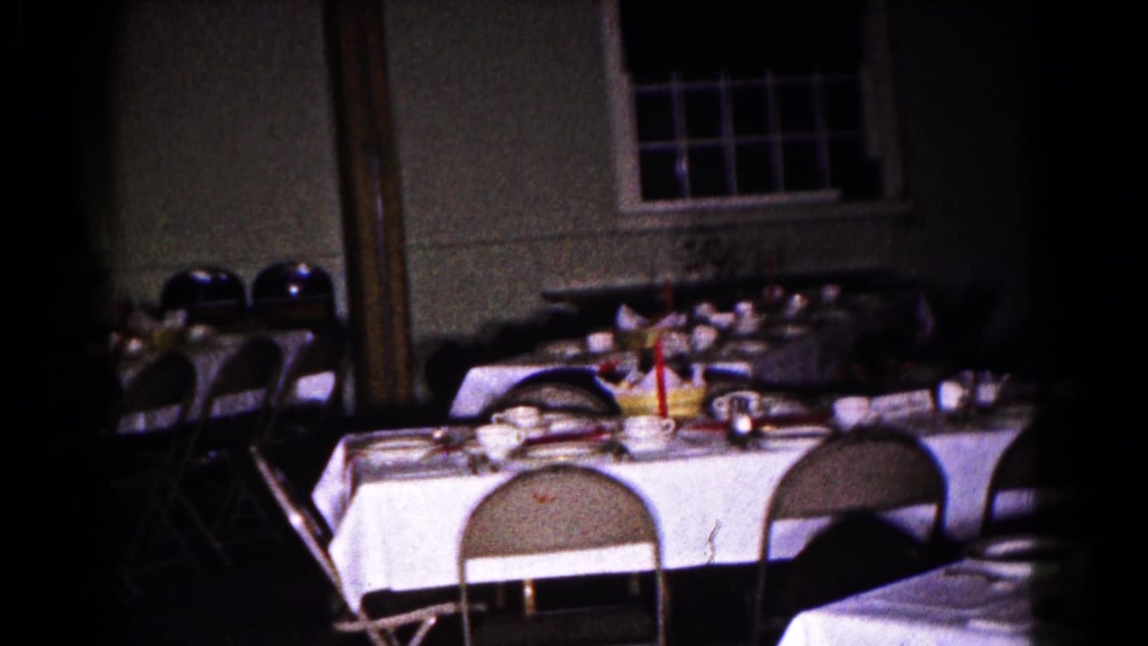 Dim banquet room with linen-covered tables, folding chairs, dishes, glassware, and a red candle centerpiece.