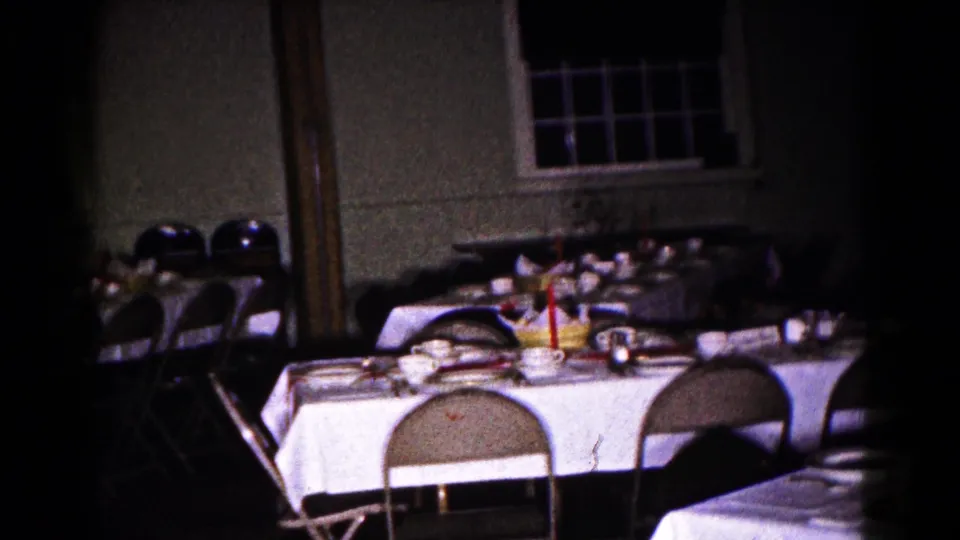 1961 Hagerstown home-movie frame showing an empty banquet room with white tablecloths, folding chairs, place settings, and dark windows.