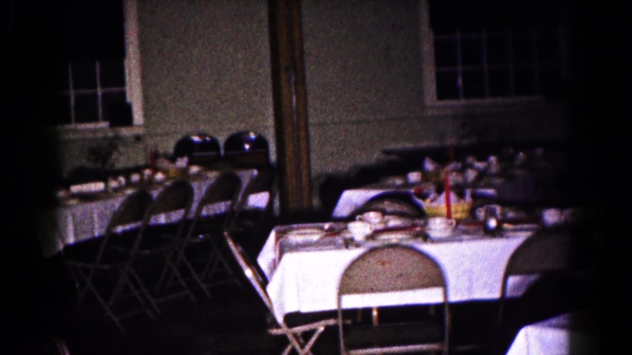 Prepared tables and folding chairs in a dark room, with dishes, cups, glassware, and windows behind them.