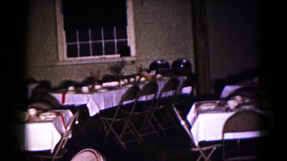 1961 8mm frame of an empty banquet or dining room in Hagerstown, with place settings, folding chairs, and a dark windowed wall.