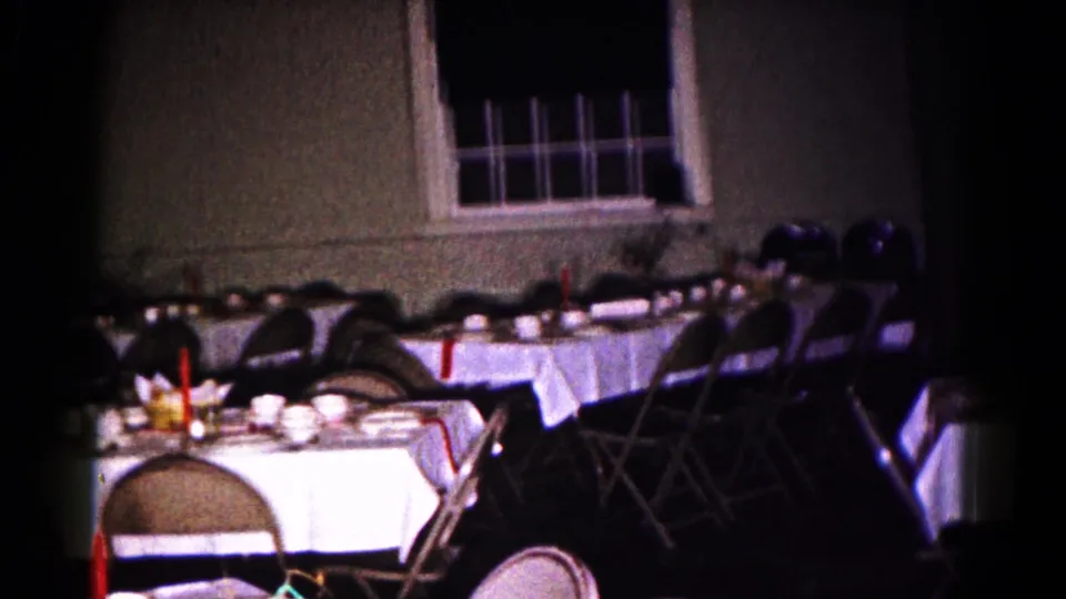 Hagerstown 1961 home-movie still showing empty linen-covered tables, folding chairs, tableware, and a red candle in a dim room.