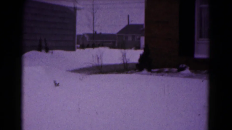1961 Hagerstown home-movie frame showing snow across a residential yard beside a brick-and-siding house.