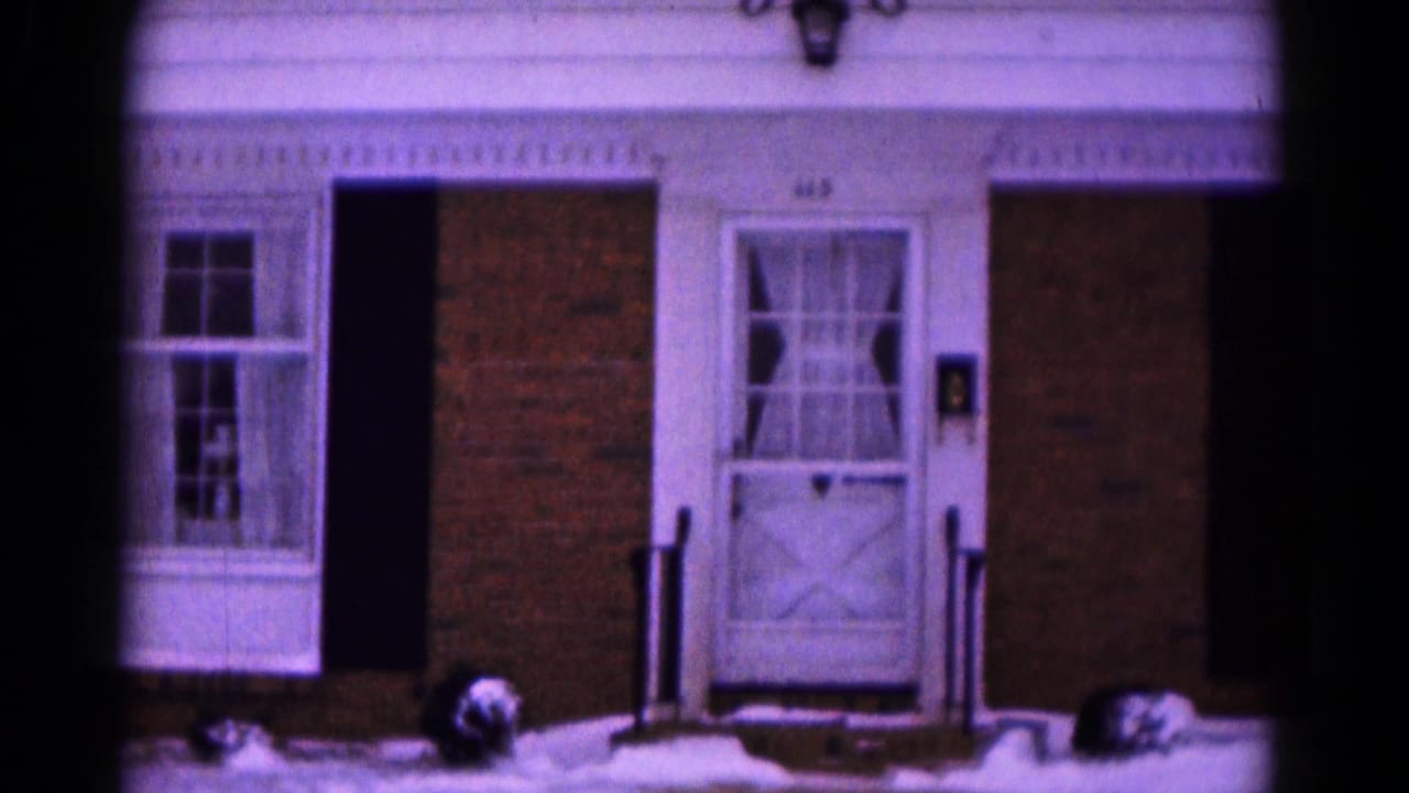 Snow-covered brick house entrance with white door, curtained windows, shutters, and porch rails.