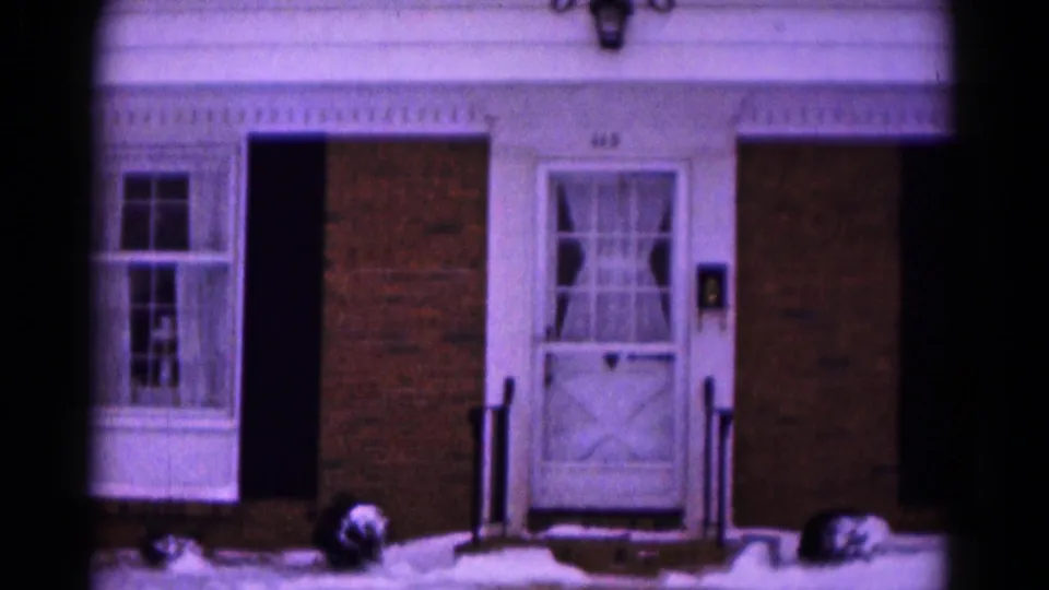 1961 Hagerstown home-movie frame showing a brick house front with white-trimmed door, curtained windows, shutters, and snow at the steps.