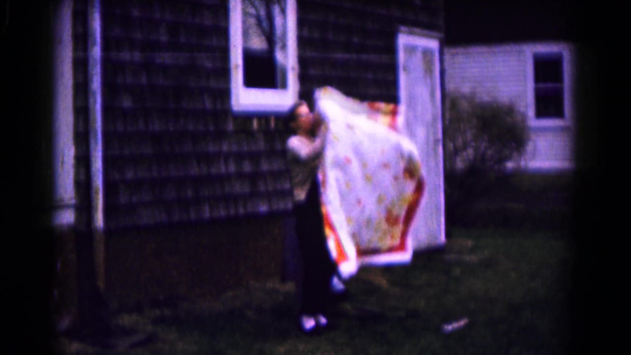 Woman lifting a patterned quilt near a clothesline outside a shingled house.