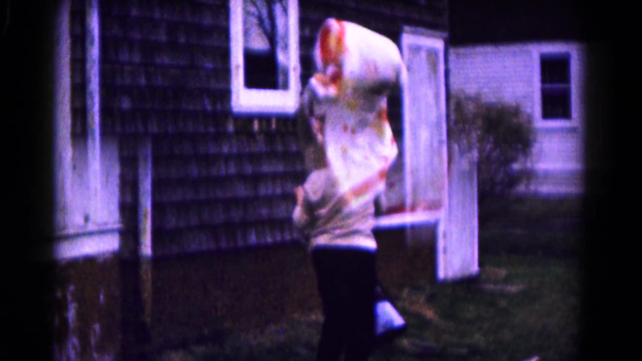 The woman stands in the yard with the quilt raised high, framed by the shingled wall, white trim, door, and grass.