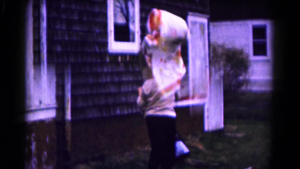 The woman stands in the yard with the quilt raised high, framed by the shingled wall, white trim, door, and grass.