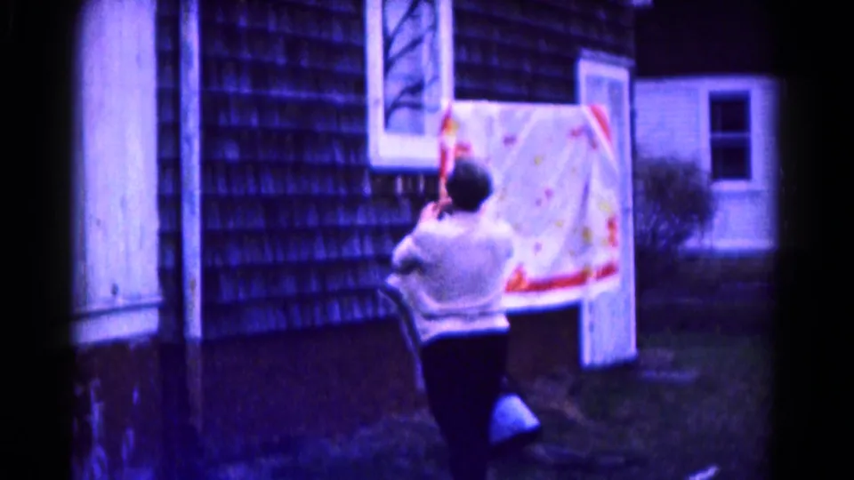 The patterned quilt hangs from the clothesline as the woman stands below, with the house wall, door, window, and yard visible.