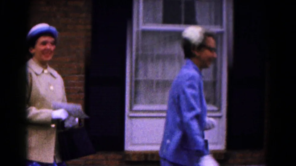 A woman in a blue suit moves across the foreground while another hat-wearing woman smiles near the brick building entrance.