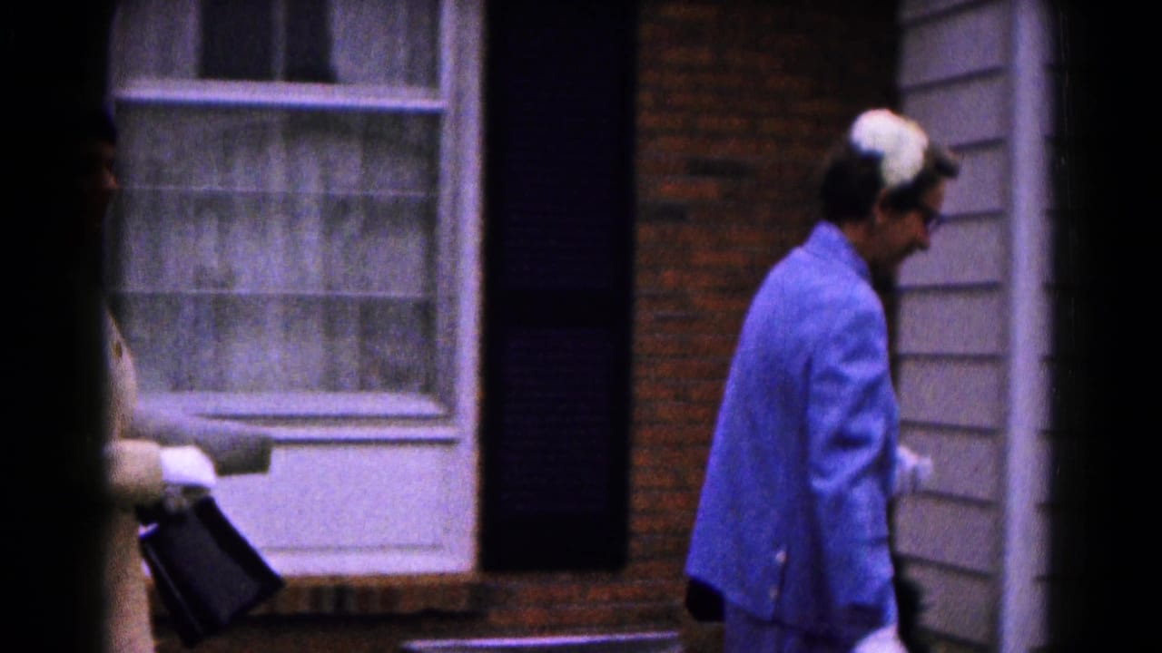 Woman in a blue coat and white gloves walks past a brick house window.