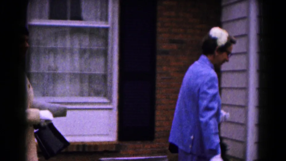 In 1961 Hagerstown home-movie footage, a woman in a blue coat and white gloves walks beside a brick house facade and curtained window.