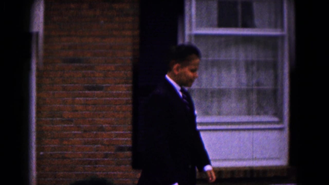 A boy in a dark suit walks past a brick house window in the 1961 Hagerstown home-movie scene.