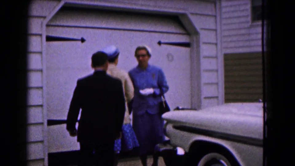 Two women and a boy in formal clothing stand near a garage door and white car in the 1961 Hagerstown home-movie clip.