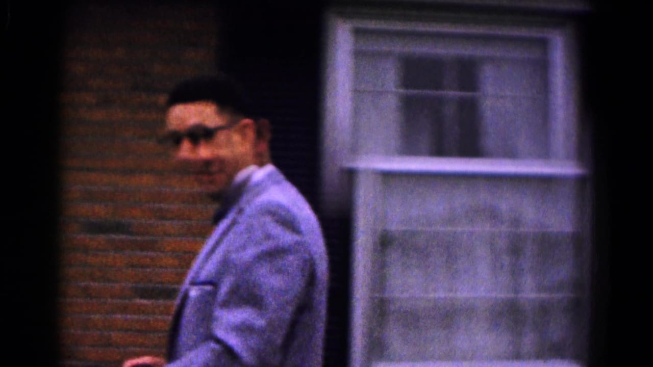 Man in a light suit and sunglasses walks past a brick house window.