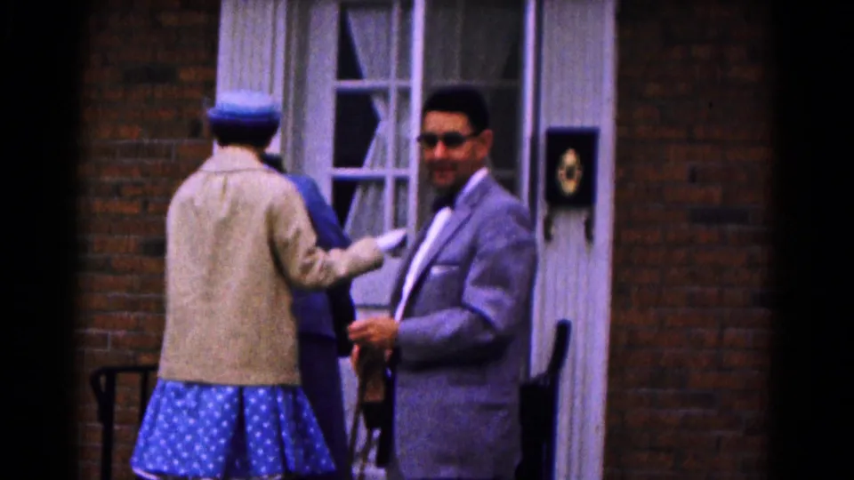 Outside the brick house, the woman faces the doorway as the suited man turns back toward the camera during the 1961 home-movie scene.