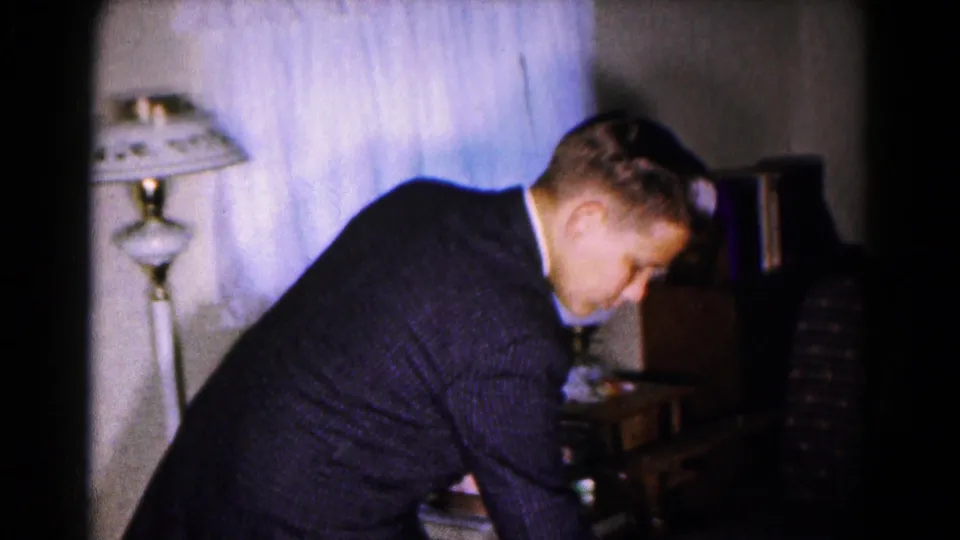 Indoors, a man in a dark suit bends near a lamp, curtains, chair, and small table in a private home interior.