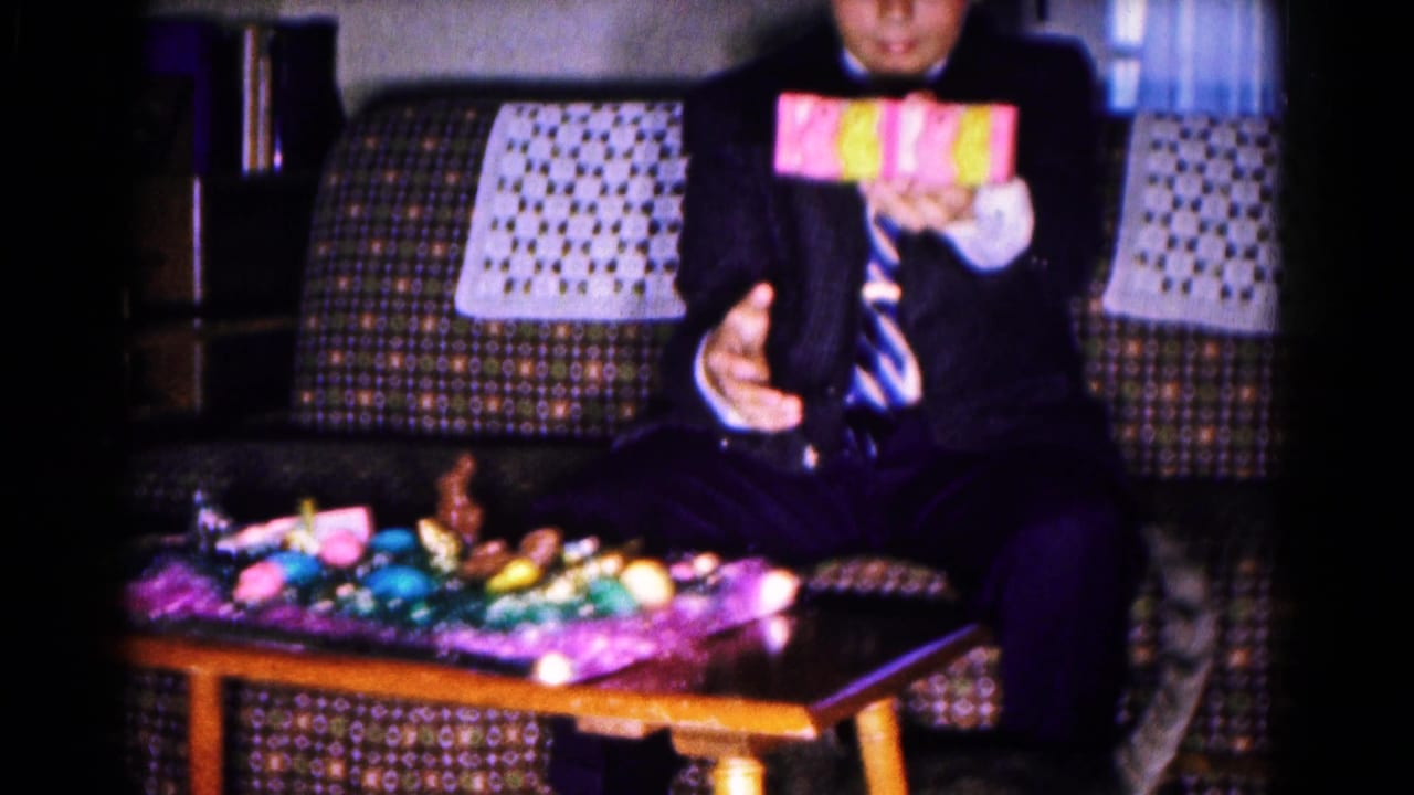 Man in a dark suit sits on a patterned couch holding a colorful wrapped gift.