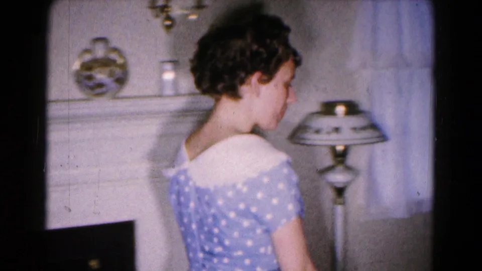 A young woman in a blue and white polka dot dress is seen from behind in a private Hagerstown living room, near a lamp and mantel.