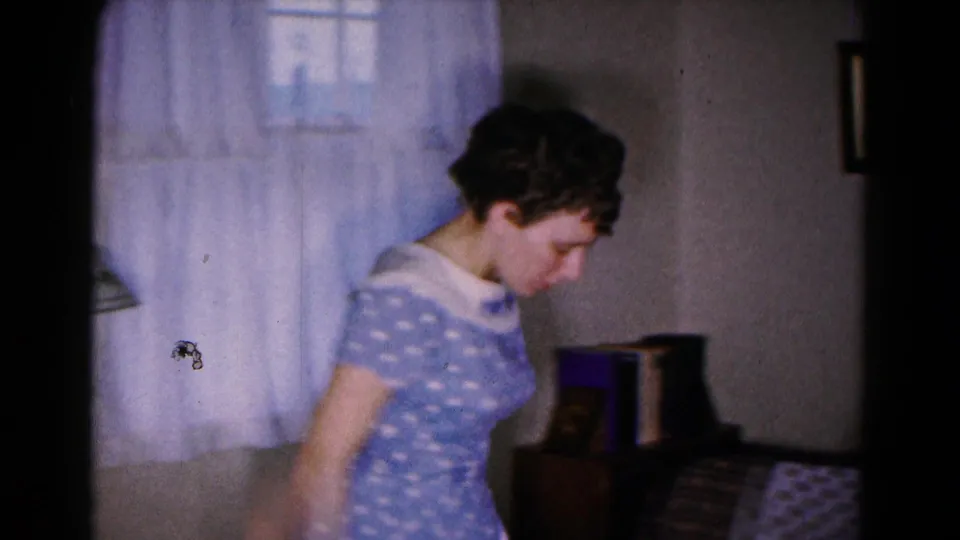 The woman in the blue polka dot dress moves through the living room beside a curtained window and small furnishings in the 8mm frame.
